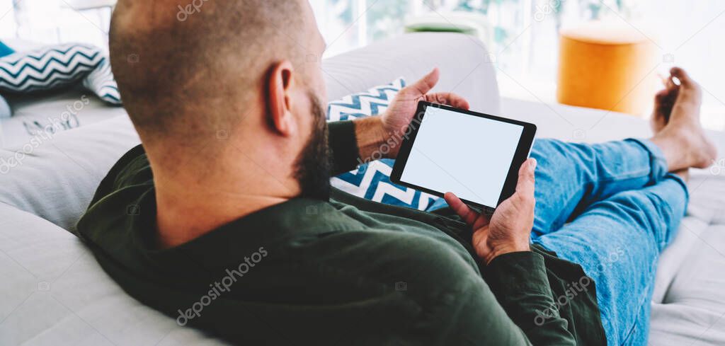 Back view of crop faceless bearded male in casual outfit sitting with outstretched legs on couch with pillows while watching video on tablet and relaxing on weekend