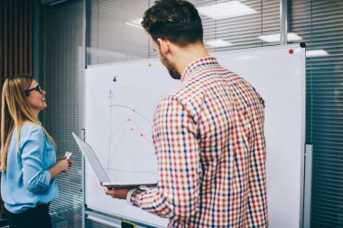 Gündelik giysiler içinde neşeli genç iş arkadaşları ofiste yeni bir projede çalışırken dizüstü bilgisayar kullanarak büyük beyaz tahtada grafikler çiziyorlar.