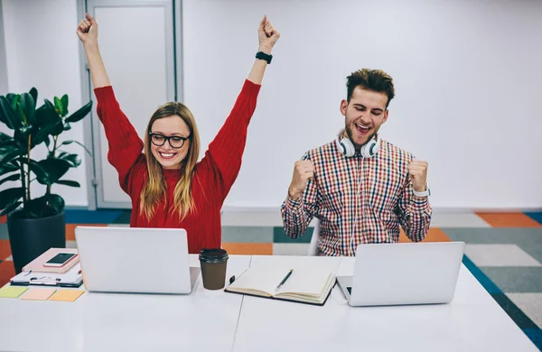 Masada dizüstü bilgisayarlarla oturup zaferi kutlarken mutlu bir şekilde gülümseyen neşeli genç iş arkadaşları.