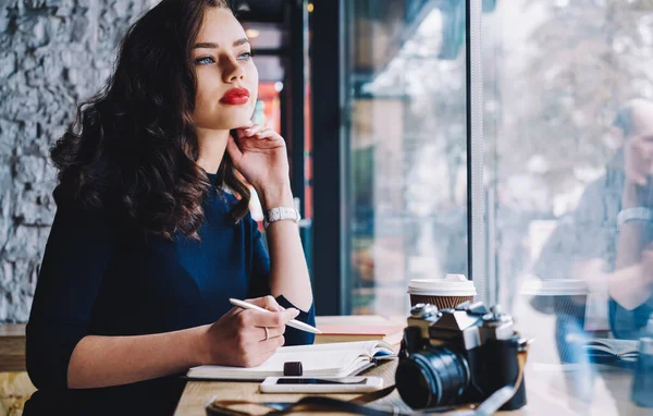 Kırmızı dudaklı, dalgın bir kadın fotoğrafçı not alırken projeyi düşünüyor ve pencerenin yanında fotoğraf makinesiyle masada oturuyor.