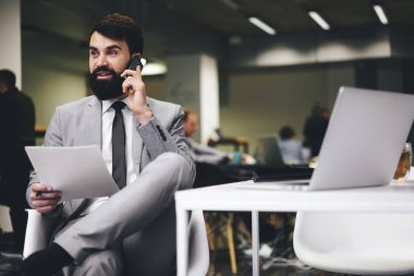 Resmi takım elbiseli düşünceli girişimci masada bacak bacak bacak üstüne atmış bir şekilde oturuyor ve iş sırasında cep telefonuyla konuşuyor.