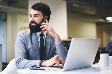 Resmi takım elbiseli dalgın sakallı girişimci modern ofisteki cep telefonuyla konuşurken masada oturup dizüstü bilgisayarını karıştırıyor.