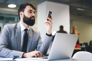 Ciddi bir girişimci resmi giysiler içinde dizüstü bilgisayarla masada oturuyor ve modern çalışma alanında yeni bir iş projesi üzerinde çalışırken akıllı telefon kullanıyor.