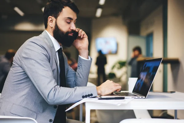 Resmi takım elbiseli, sakallı, netbook 'la masada oturan ve ofiste çalışırken cep telefonuyla konuşan çalışanın yan görüntüsü.
