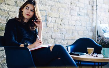 Kafe sandalyesinde elinde not defteri ve kahveyle oturan ve not defterine notlar yazarken telefonla konuşan ve başını çeviren günlük kıyafetler giyen genç bir kadın serbest yazar.