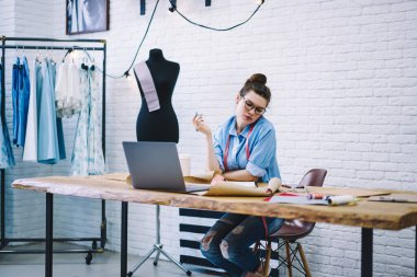Düşünceli bayan moda tasarımcısı, yeni koleksiyon için eskizler çiziyor. Stüdyoda dizüstü bilgisayarla, mankenlerle ve askılarda sıra sıra elbiseler ile oturuyor.