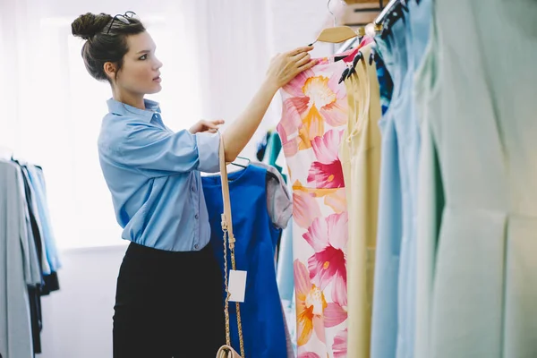 Mavi gömlekli, gözlüklü zarif bayanın modern atölyede rengarenk giysileri seçişi.