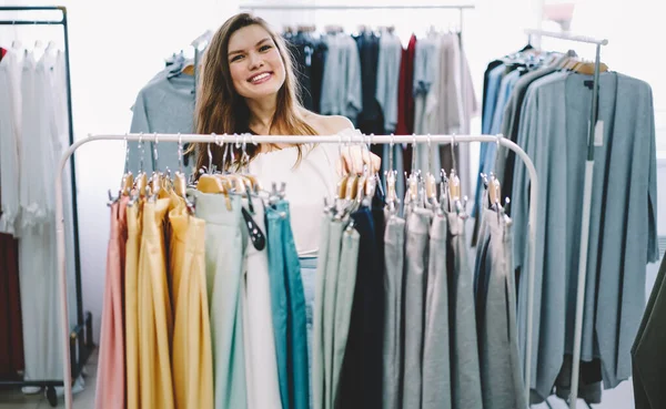 モダンな店内で新しいアパレルを選んでカメラを見ながら 服の棚の後ろに立つ幸せな女性の買い物客 — ストック写真
