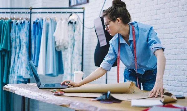 Konsantre kadın terzi gözlüklü, dizüstü bilgisayarda gezerken aynı zamanda giysili modern atölyede üzerinde kağıt olan bir tasarım projesi yaratıyor.