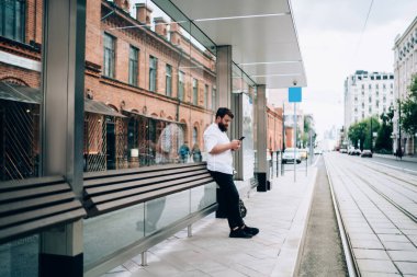 Kenar mahalledeki tramvay istasyonunun kaldırımında dikilirken günlük kıyafet giyen, cep telefonu takan kendine güvenen sakallı adamın yan görüntüsü.