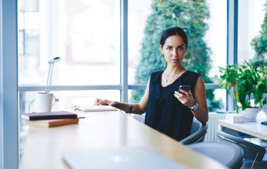 Yakışıklı, kumandalı, siyah saçlı, akıllı telefonu iş için kullanan, elinde not defterleri olan ve modern iş yerindeki kameralara bakan bir kadın.