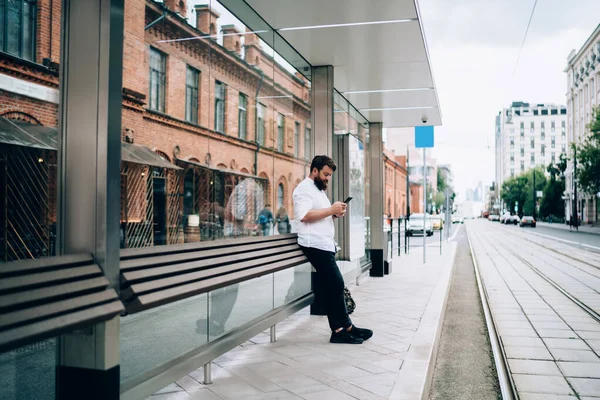 Kenar mahalledeki tramvay istasyonunun kaldırımında dikilirken günlük kıyafet giyen, cep telefonu takan kendine güvenen sakallı adamın yan görüntüsü.