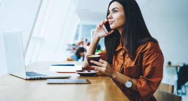 Siyah saçlı, sıradan giyinmiş, telefonla konuşan ve bir fincan kahveyle masada otururken pencereye bakan genç bir kadının yan görüntüsü.