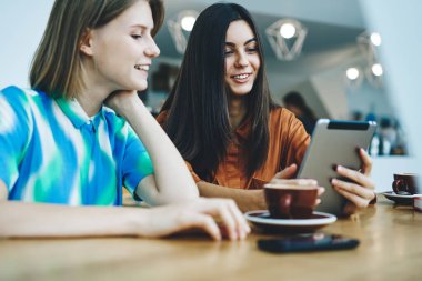 Neşeli çoklu etnik gruptan genç bayan arkadaşlar tabletten komik videolar izliyorlar modern kafeteryada fincanlar ve akıllı telefonlar ile masada oturuyorlar.