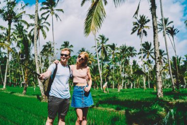 Günlük kıyafetleri ve güneş gözlükleriyle mutlu genç adam ve kadın tropikal bahçede yürüyor ve selfie çubuğuyla gülümsüyor.