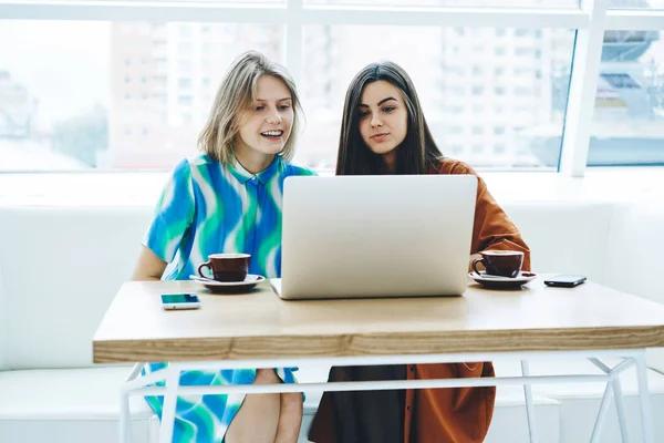 穿着休闲装 拿着咖啡和智能手机坐在桌旁的积极女学生 同时在大学的笔记本电脑上一起工作 — 图库照片