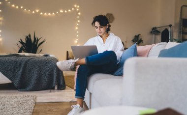 Rahat giysiler içinde, kulaklık takmış, modern netbook 'a takmış, evinde rahat bir kanepede bacak bacak bacak bacak üstüne atmış genç bir esmer.