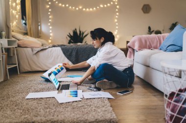 Yan görünüş, günlük kıyafetler giyen, evrak işleri yapan ve rahat yatak odasında bacak bacak bacak üstüne atmış bir şekilde otururken modern netbook okuyan çekici bir kadın.