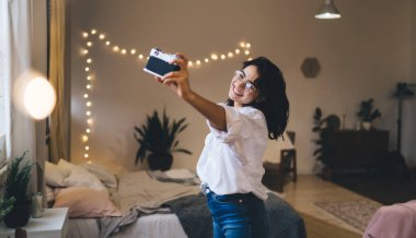 Saçları dökülmüş, eski moda fotoğraf makinesiyle rahat bir odada duvarda parlayan ışıklarla kendi portresini çeken hoş bir kadın.