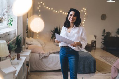 Elinde belgeyle, yatak odasında dekorasyonlu bir şekilde duvarda ışıklandırmayla kameraya bakan olumlu bir kadın.