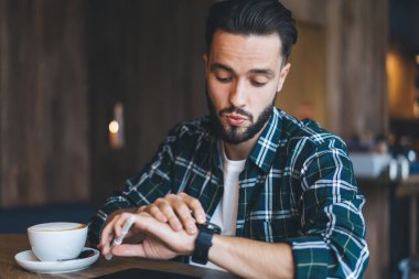 Kafeterya masasında oturmuş, takılabilir bilek saatini kontrol eden beyaz tenli bir erkek fazla mesai yapmaktan etkilenmiş hippi bir adamın dokunmatik akıllı adam ekranına bakmasından etkilenmiş.