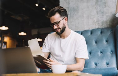 Bilgi çalışması ve sınav hazırlığı için bilgi notları okurken gülümseyen erkek öğrenci, 20 'li yaşlarda yetenekli bir adam dizüstü bilgisayarla birlikte çalışma alanında oturmuş ders kitabı bilgilerini analiz ediyor.