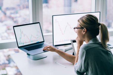 Gözlüklü ve gömlekli genç bir kadının modern ofisteki dizüstü bilgisayarında istatistiksel bilgilerle çalışırken yan görünümü