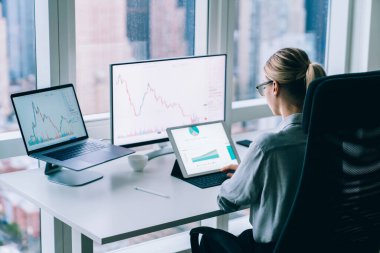 Tanımlanamayan kadın yöneticinin resmi kıyafetleriyle tabletle çalışmasının ve pencere kenarındaki cihazların arasında ofis sandalyesinde çizelgeleri analiz etmesinin arka planı.