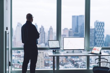 Cep telefonuyla pencereden ve masadan ekrandaki grafikleri temsil eden bilgisayarlarla bakan isimsiz erkek girişimcinin arka planı