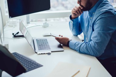 İş yerinde bilgisayar ve dizüstü bilgisayarla çalışırken cep telefonunda mesajları okurken gömlekli ve gözlüklü, düşünceli, düşünceli bir erkek yöneticinin yüksek açılı yan görüntüsü.