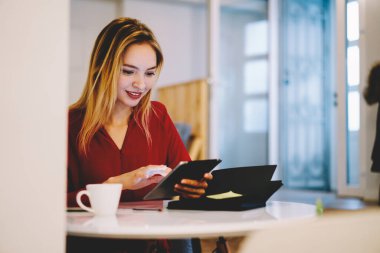 Gündüz vakti hafif ferah bir kafede bir bardak içkiyle masada otururken günlük kıyafetlerle tablete yazı yazan odaklanmış bir öğrenci.