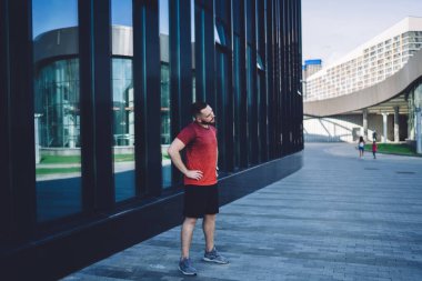 Spor kıyafetli bir sporcunun modern camdan yapılma bir binanın yanındaki kaldırımda dikilirken ısınma egzersizleri yapması.