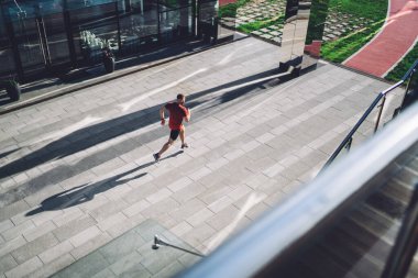 Yukarıdan bakıldığında, kırmızı tişörtlü, spor yapan, koşan ve bakışlarını başka yöne çeviren, tanınmayan genç bir atletizm sporcusu görülüyor.
