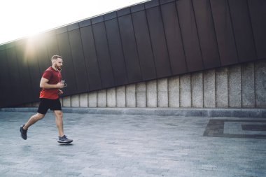Kulaklıklı, elinde telefonla caddede koşan ve sabah antrenman yaparken müzik dinleyen kulaklıklı erkek sporcunun yan görüntüsü.