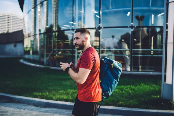Ciddi sakallı, spor kıyafetli, akıllı telefon ve sırt çantalı bir adamın modern cam binanın yanında yürürken yüzünü çevirmesi.