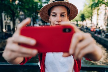 Gülümseyen genç bayan gezgin şapka takıyor ve günlük giysiler giyiyor cep telefonuyla selfie çekiyor Amsterdam kanalının üstündeki köprüde duruyor.