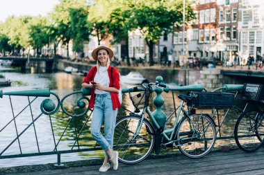 Amsterdam 'da köprü korkuluklarına yaslanırken cep telefonuyla sosyal medyada gezinen mutlu genç bir kadın gezginin tüm vücudu.