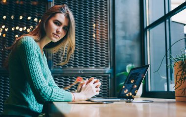 Kendine güvenen, uzun saçlı, yeşil kazaklı çekici bir kadın. Ahşap masadaki modern rahat restoranda oturuyor ve kameraya bakıyor.