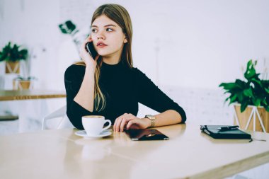 Düşünceli, uzun saçlı, siyah kazaklı bir kadın akıllı telefondan konuşuyor. Bir fincan sıcak içecek içiyor ve rahat bir kafede başka yere bakıyor.