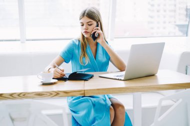 Genç bayan sekreter akıllı telefondan konuşuyor ve not defterine talimatlar yazıyor. Masada dizüstü bilgisayarı ve sıcak içeceklerle oturuyor.