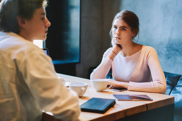 Young serious female watching at male friend while sitting in cafe in daytime and drinking cup of drink and wearing casual outfit with tablet at table
