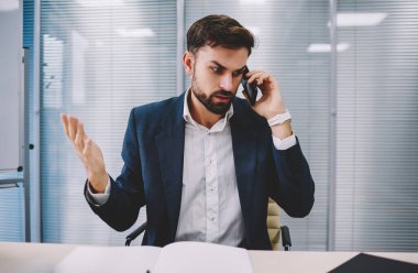 Kızgın iş adamı resmi mavi ceket ve gömlek giyer masada oturur ve cam duvara karşı çağdaş çalışma alanında akıllı telefonla konuşur.