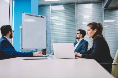 Resmi giysiler içinde bir grup düşünceli iş arkadaşı konferans masasında oturmuş modern ofiste projenin detaylarını tartışıyorlar.