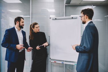 Resmi kıyafetler içinde bir grup iş arkadaşı boş tahtanın yanında duruyor ve modern ofiste şirket stratejisi hakkında tartışıyorlar.