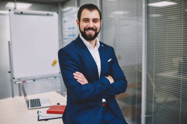 Resmi takım elbiseli, göğsünde çapraz kollar olan, modern ofiste dizüstü bilgisayarla çalışırken dişlek gülümsemesi olan ve kameraya bakan tatmin olmuş bir adam.