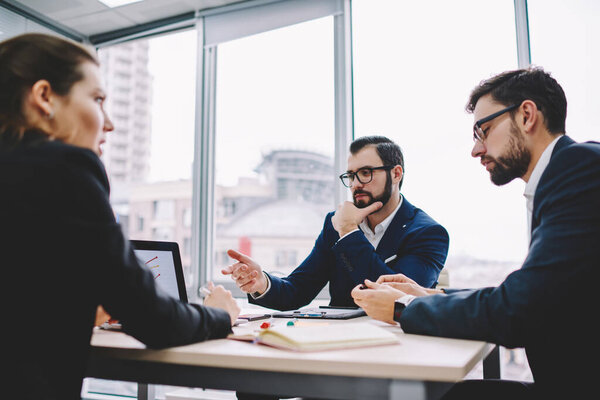 Thoughtful business people in formal clothes sitting at table and using laptop with presentation while working on project in modern workspace