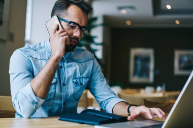 Ciddi erkek serbest çalışan, günlük giysiler içinde masada oturmuş, akıllı telefondan sohbet ediyor ve kafede çalışırken dizüstü bilgisayarını karıştırıyor.