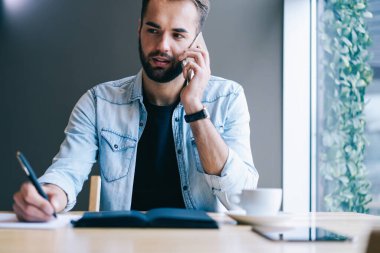 Günlük giysiler içinde sakallı bir öğrenci bir bardak içki ve defterle masada oturuyor ve hafif çalışma alanında cep telefonuyla konuşurken kâğıt kağıtlara not alıyor.