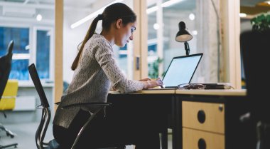 Üzerinde gündelik giysiler olan, siyah saçlı, masa başında oturan ve modern çalışma ortamında netbook 'a yazan kadının yan görüntüsü.