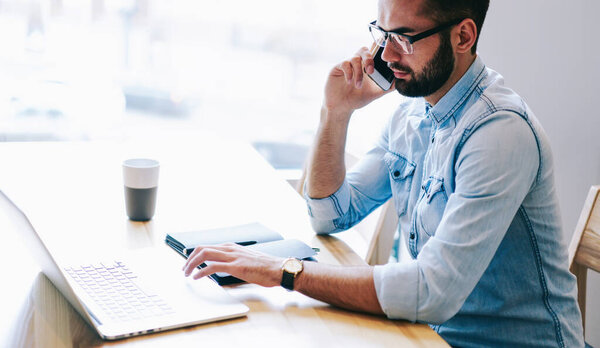 Side view of concentrated male freelancer sitting at table with notepad and cup of hot drink while using laptop and having conversation on smartphone